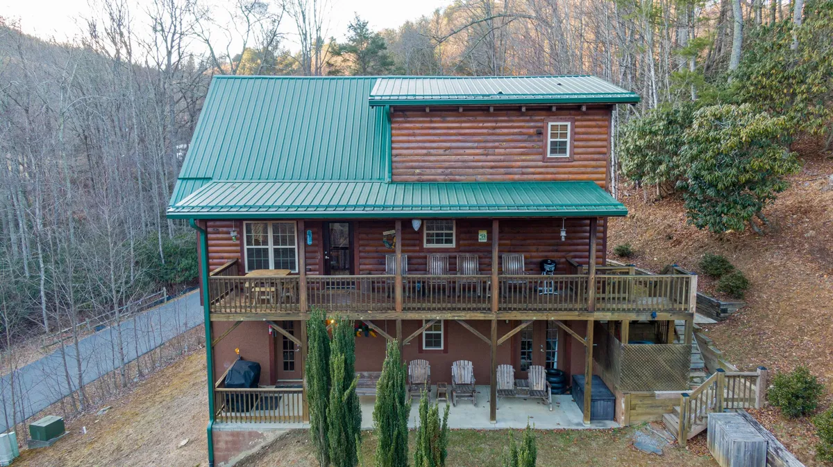 This image shows the log cabin's exterior with a green metal roof, a welcoming front porch furnished with seating, and lush trees all around. The porch is clearly set up for relaxing and enjoying the view, while the setting hints at both sunrise coffee moments and sunset gatherings.