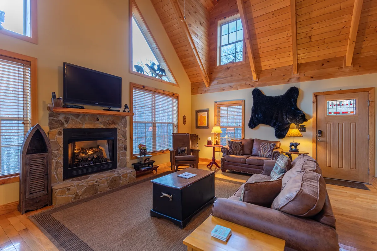 A warmly lit main living area featuring a rustic fireplace, large windows that offer glimpses of the outdoors, exposed wooden beams, a comfortable sofa set, and a coffee table. The scene radiates coziness and invites relaxation, signaling a perfect space for families (including pets) to unwind after outdoor adventures.
