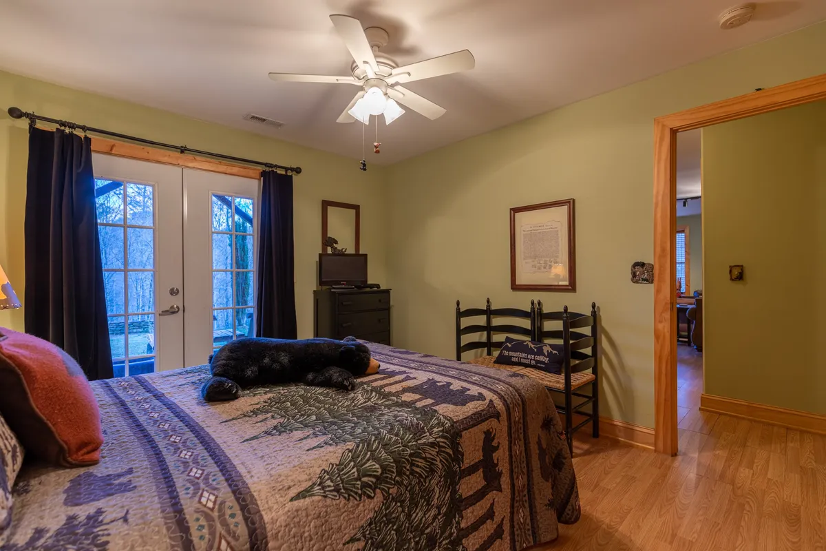 A welcoming bedroom featuring a bed with a patterned quilt, double French doors letting in natural light, and a simple nightstand with a lamp. The space feels open and restful, suggesting privacy and comfort.