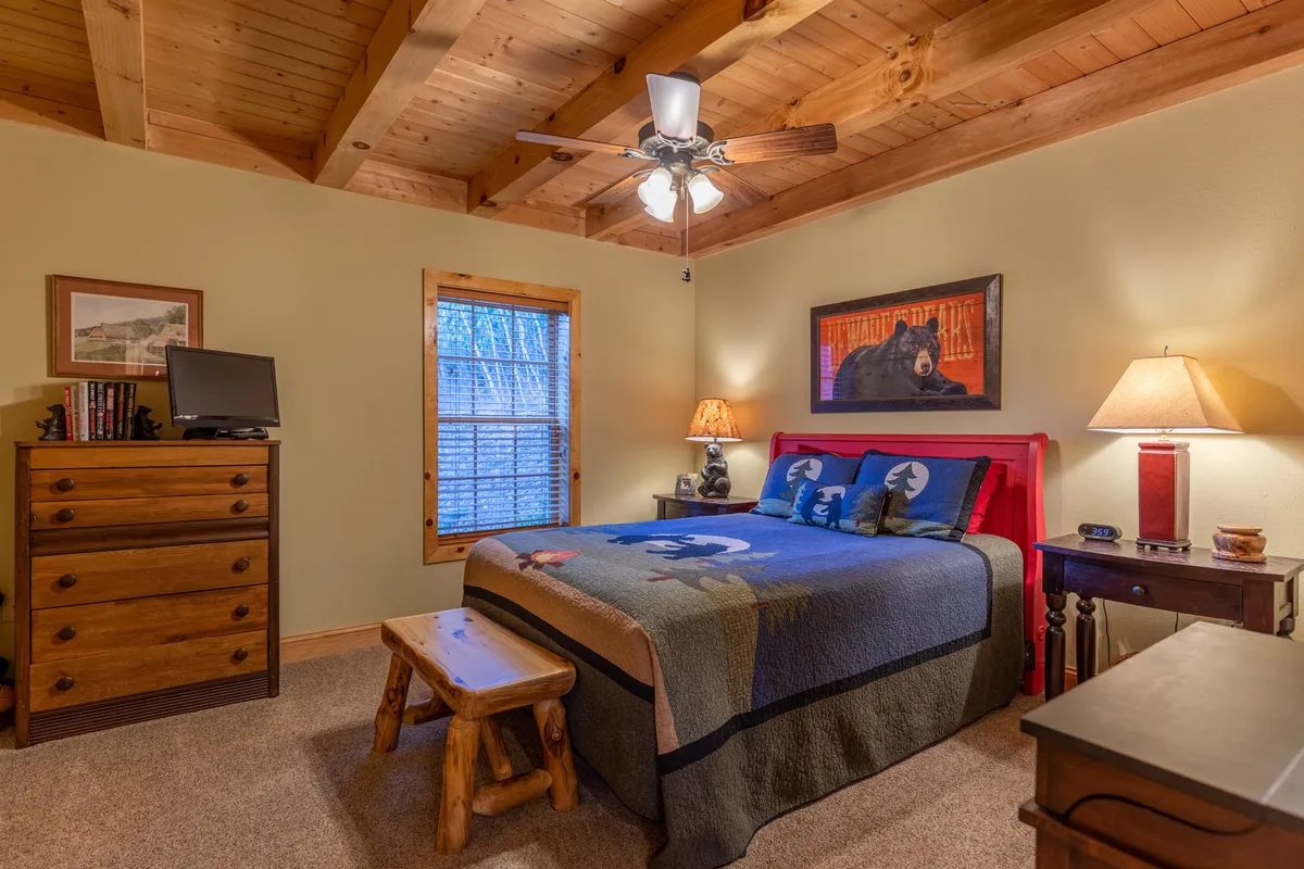 A cozy bedroom with a decorative blanket on the bed, framed bear art above the headboard, wooden furniture, and a lamp softly illuminating the room. The window invites gentle daylight into the restful space.