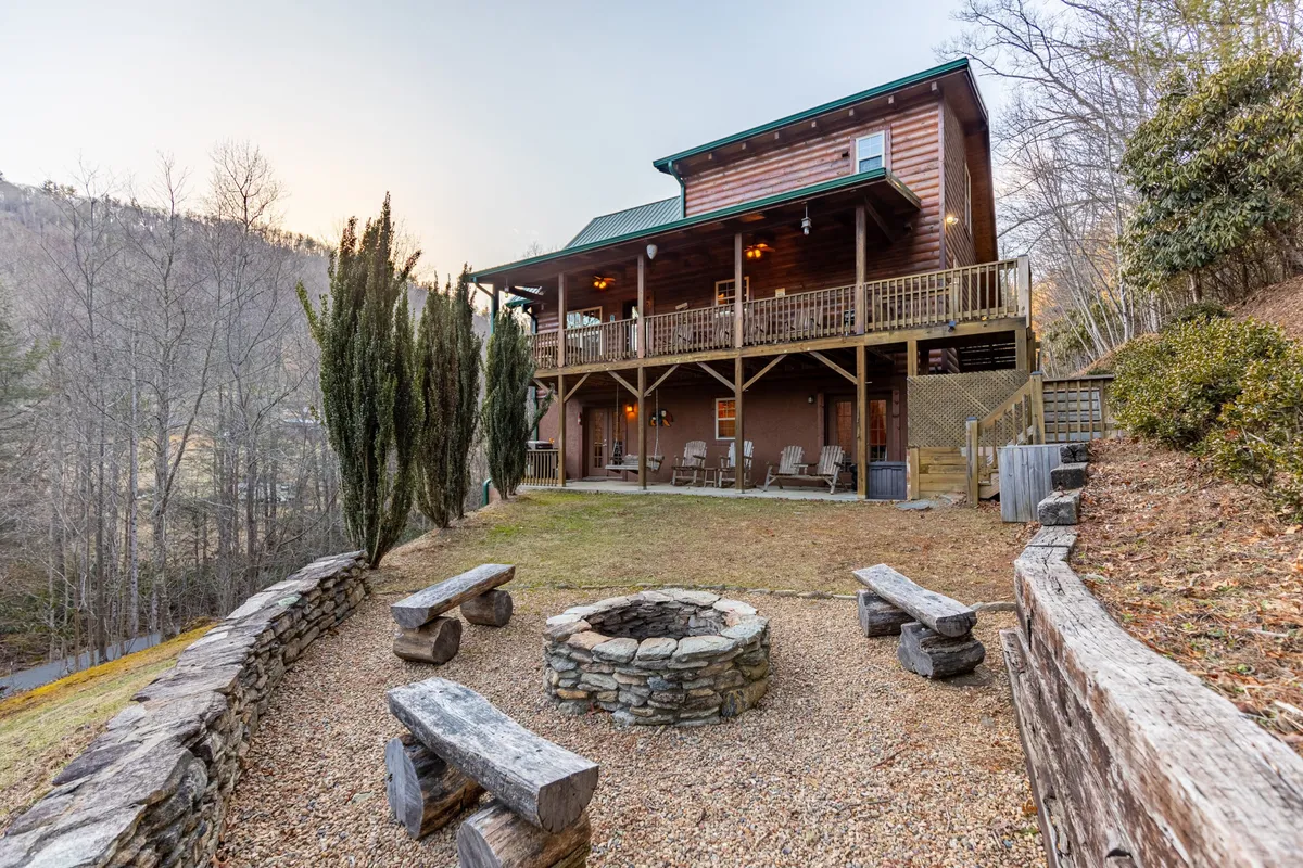 The image features the inviting exterior of Bear Creek cabin at dusk, with a cozy fire pit surrounded by comfortable seating in the foreground, perfect for gathering with friends and family. The cabin's charming facade glows warmly, suggesting a welcoming atmosphere ideal for relaxed evenings under the stars.