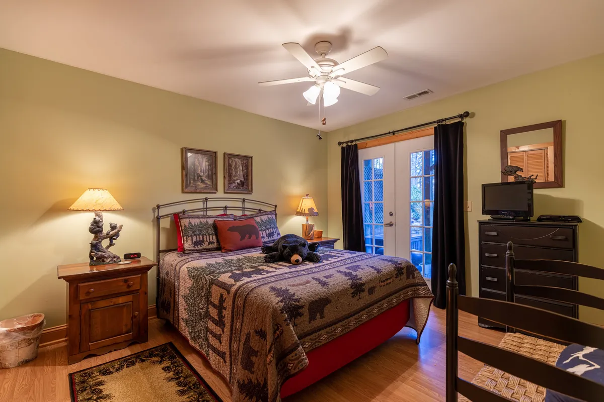 A cozy bedroom scene with a bed draped in a patterned blanket, a nightstand and lamp, a black bear plush toy, and double doors leading directly outside. The room is bright and inviting, offering both restful sleep and immediate access to the outdoors for morning walks or pet outings.