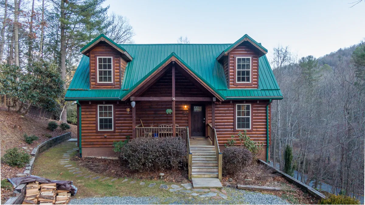 This image showcases the exterior of the Bear Creek log cabin with its charming green metal roof, wooden steps leading to the entrance, and the surrounding trees that create a cozy, inviting setting. The porch and yard suggest spaces for outdoor fun and relaxation, such as gathering for campfires or nature walks.