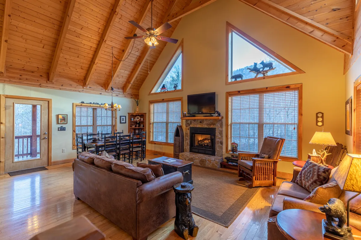 An expansive interior with large windows framing mountain views, a stone fireplace, a wooden dining table with chairs, and a plush leather couch. The natural light and spacious layout reflect both comfort and a connection to the scenic outdoors, reinforcing the mountain retreat vibe.